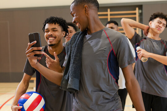 Diverse male volleyball partners interacting over smartphone in school gym with ball and towel