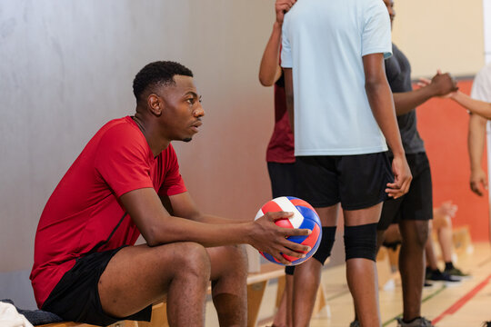 Diverse male volleyball teammates gathering on bench sideline court holding volleyball inside gymnas