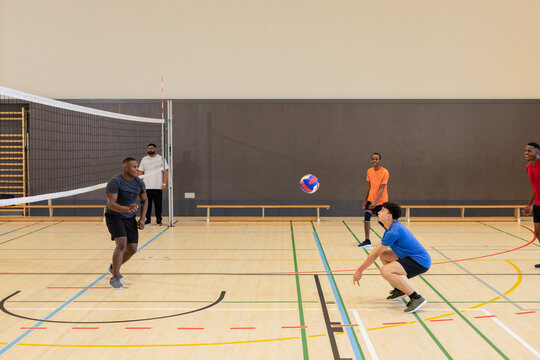 Diverse male players playing volleyball inside gymnasium court with volleyball net and wooden benche