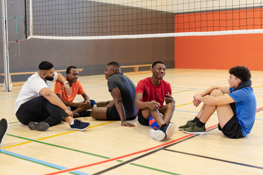 Diverse male teammates in sportswear sitting on volleyball gymnasium chatting around net and volleyb
