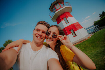 Smiling couple taking a selfie in front of a red and white striped lighthouse on a sunny day, symbolizing love, travel, summer vacation, and joyful outdoor adventure.