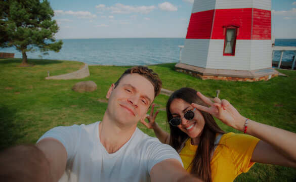 Happy couple taking a cheerful selfie near a red and white lighthouse by the sea, both smiling and showing peace signs, enjoying summer vacation and seaside fun.