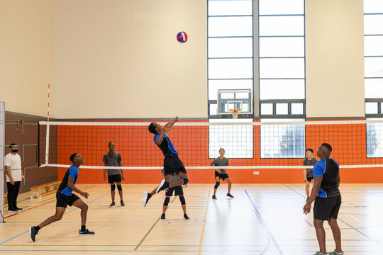 Asian male volleyball player jumping near net spiking volleyball in gym under windows