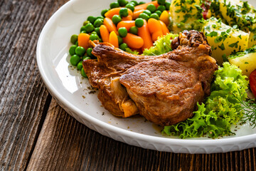 Baked veal cutlet with bone, boiled potatoes, mini carrots and green peas on wooden table