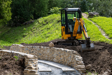 Yellow excavator working on soil near stone wall, symbol of construction, land development, heavy machinery, building industry and engineering project outdoors