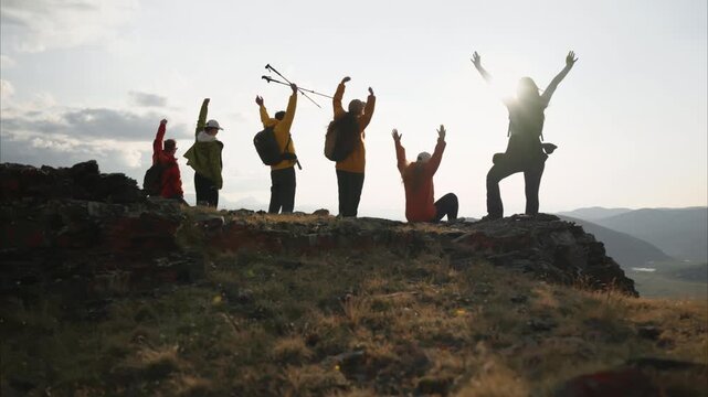 Wide angle bottom view of a group of diverse hikers man and woman celebrating reaching the summit in the Altai Mountains. Trekkers raise their hands in the background of sunset sky