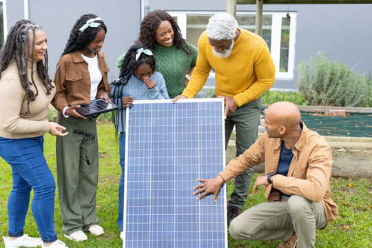 Diverse family with solar technician examining large solar panel and small module on grass lawn