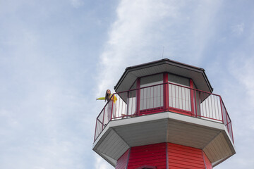Young woman in yellow shirt stands on the balcony of a red and white lighthouse, pointing outwards with a smile, under a bright blue sky with scattered clouds.