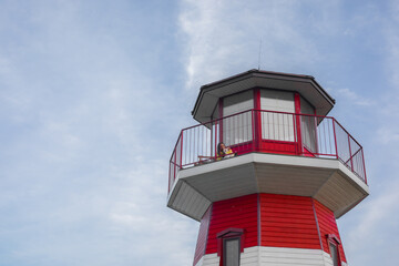Young woman in a yellow shirt with a white dog on the balcony of a red and white lighthouse, overlooking the sea under a clear blue sky.