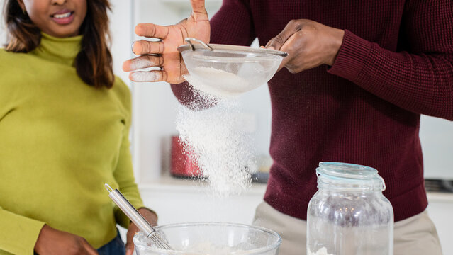 Indian woman mixing bowl and whisk, African American man sifting flour in sieve on kitchen counter