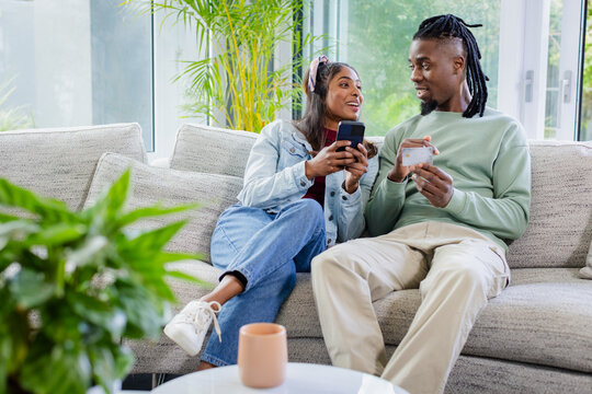 Diverse couple sitting on light gray sofa in home living room holding smartphone and credit card