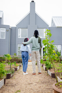 Diverse couple embracing on gravel pathway flanked by wooden raised beds with plants near gray home