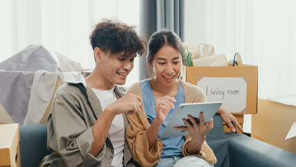Young happy Asia couple sit on sofa couch use digital tablet online shopping furniture from decoration store in living room at home. Husband wife move new house, Apartment Rent And Ownership Concept.