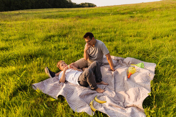 A father and his young son relax on a soft blanket in a lush, green field. They share a joyful moment filled with laughter and snacks on a warm, sunny day
