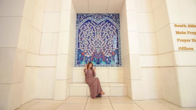 A young woman in a modest dress sits in front of a decorative blue tile mosaic in Sheikh Zayed Mosque, Abu Dhabi, United Arab Emirates