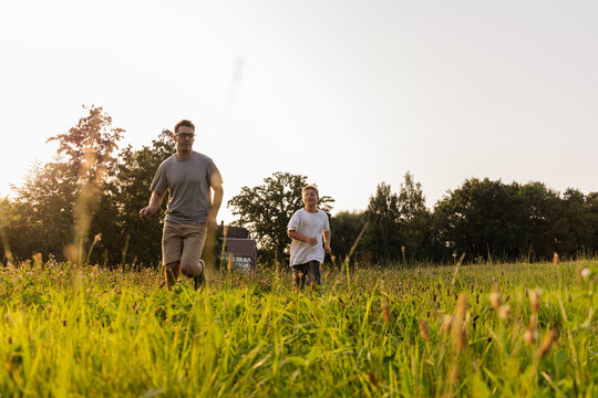 Father and son share joyful moments as they run through a lush green field during the late afternoon. The sun sets behind the trees, creating a warm and inviting atmosphere