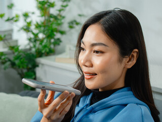 Young asian Casual woman using AI Voice Assistant App on Mobile phone, Talking to Chatbot Job Consulting, Remote Work and AI Assistant for Business Sitting on Couch in Living room at home.