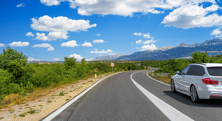 A white car drives along a winding mountain road surrounded by greenery and scenic views under a...