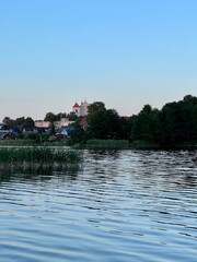 Obraz premium Tranquil rural scene featuring a lake at sunset with a soft sky. The foreground shows tall grass and water plants by the lakeside. Across the lake, a church with a pink facade and multiple spires 