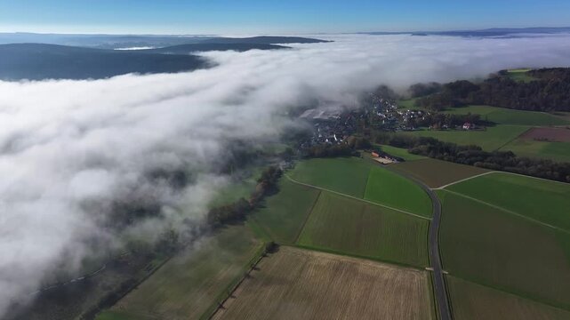 Aerial view of a thick blanket of fog partially obscures a quaint village nestled amidst vibrant green fields, Eichenzell, Hessen, Germany.