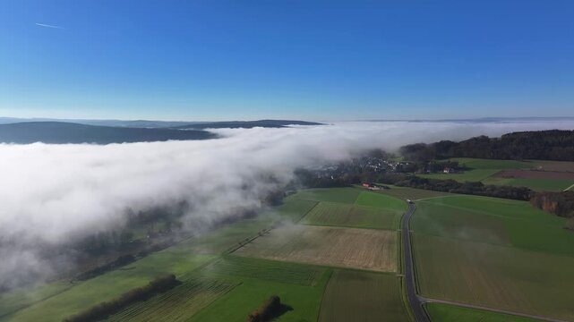 Aerial view of the lush green fields and a blanket of fog partially covering the town of Eichenzell, creating a serene contrast, Eichenzell, Hessen, Germany.