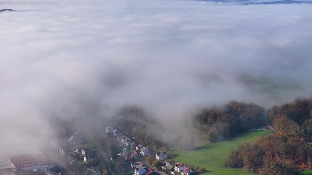 Aerial view of a thick blanket of clouds hovering over a town nestled among green fields and forests, creating a scene of ethereal beauty, Eichenzell, Hessen, Germany.