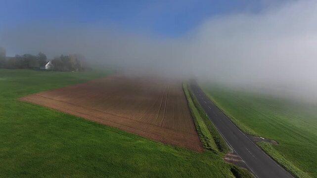 Aerial view of a road cutting through fields, partially obscured by a dense fog, creating a mysterious atmosphere, Eichenzell, Hessen, Germany.
