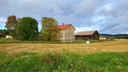A yellow wooden house stands beside a large barn on a field with ripe crops. Trees show autumn colors under a partly cloudy sky, creating a peaceful rural scene.