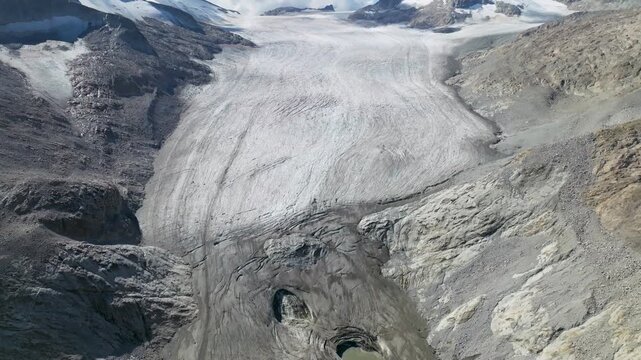 Aerial view of a glacier winding through rugged mountains, their peaks capped with snow under the sun, Brescia, BS, Italia.
