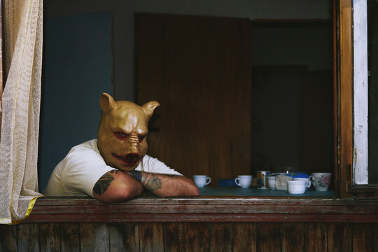 Man in pig mask leaning on windowsill with cups and tattooed arm indoors