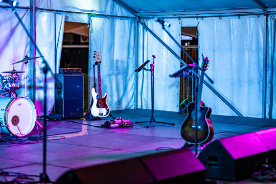A stage is set up with musical instruments including a drum kit, bass guitar, and acoustic guitar, along with amplifiers, a pedalboard, and microphones, all illuminated by vibrant blue and purple stag