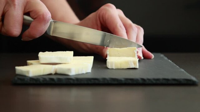 woman&rsquo;s hands carefully slicing soft brie cheese on a dark background