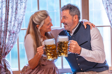 Festive couple wearing Bavarian costumes clinking beer mugs at Oktoberfest enjoying traditional...