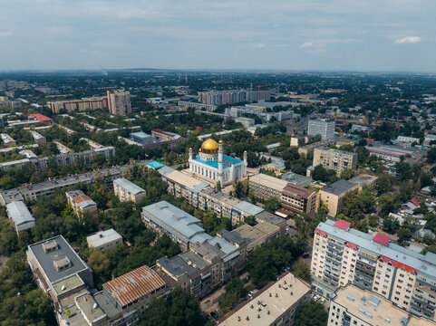 Aerial view of the grand Central Mosque's white facade and golden dome rise amidst the city's green canopy, a striking contrast under a cloudy sky, Almaty, Almaty Region, Kazakhstan.
