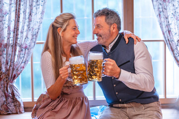 Happy couple dressed in Bavarian costumes clinking beer mugs at Oktoberfest enjoying traditional...