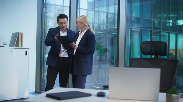 Focused Asian male in blazer discussing document with smiling blonde female standing beside him. Reviewing report or clarifying project details. Collaborating near window in modern office.
