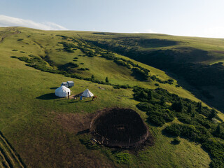 Aerial view of yurts nestled on the vibrant green hills of Assy Plateau, a circular patch of dark earth contrasting against the bright landscape, Assy Plateau, Almaty Region, Kazakhstan.
