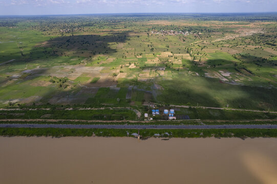 Aerial view of a road running parallel to a muddy river, with fields and buildings in the background under a cloudy sky, Babanruga, Kano, Nigeria.