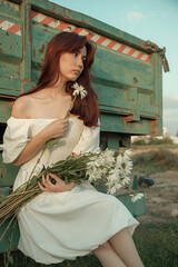 Thoughtful young woman with daisies sitting by farm trailer.Retro grain effect
