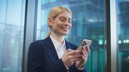 Charming blonde Caucasian woman in business suit smiling while tapping on smartphone near window. Reading positive message or checking purchase confirmation. Enjoying digital moment during workday. - Powered by Adobe