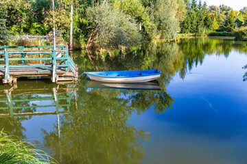 Small blue boat moored to a wooden pier on a calm lake surrounded by green trees. 
Small blue boat moored to a wooden pier on a calm lake surrounded by green trees.