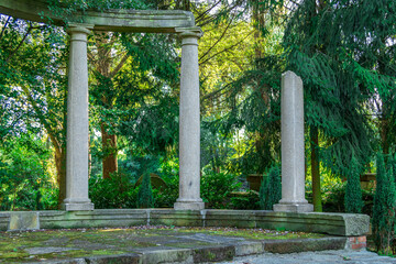 Ancient stone columns in a lush green park. Ruins of a classical structure surrounded by trees and...