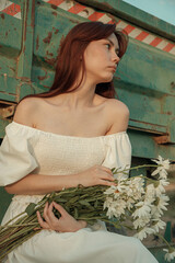 Thoughtful young woman with daisies sitting by farm trailer.Retro grain effect