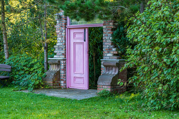 Pink door in a brick frame standing in a green garden. Mysterious entrance surrounded by trees and...