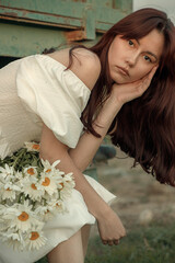 Thoughtful young woman with daisies sitting by farm trailer.Retro grain effect