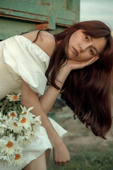 Thoughtful young woman with daisies sitting by farm trailer.Retro grain effect