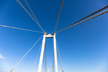 Fototapeta premium Blue Sky Suspension Bridge View From Below With Cables and White Tower Under Clear Weather. Engineering, travel, and openness across the span.