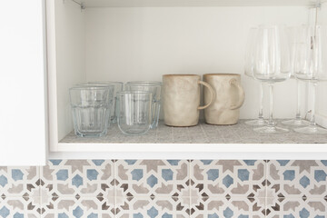 Kitchen shelf with glassware and ceramic mugs. Drinking glasses, ceramic coffee mugs, and wine glasses. Home organization