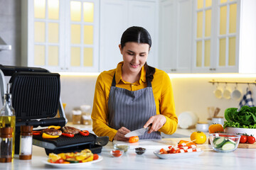 Woman cutting pepper while cooking meat and vegetables on electric grill in kitchen