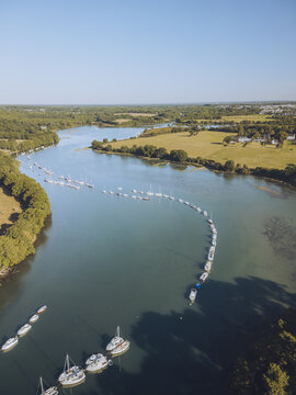 Aerial view of boats moored along the riverbank, their white hulls gleaming against the dark water, bordered by green trees and fields, Presqu'&Atilde;&reg;le de Conleau, Bretagne, France.